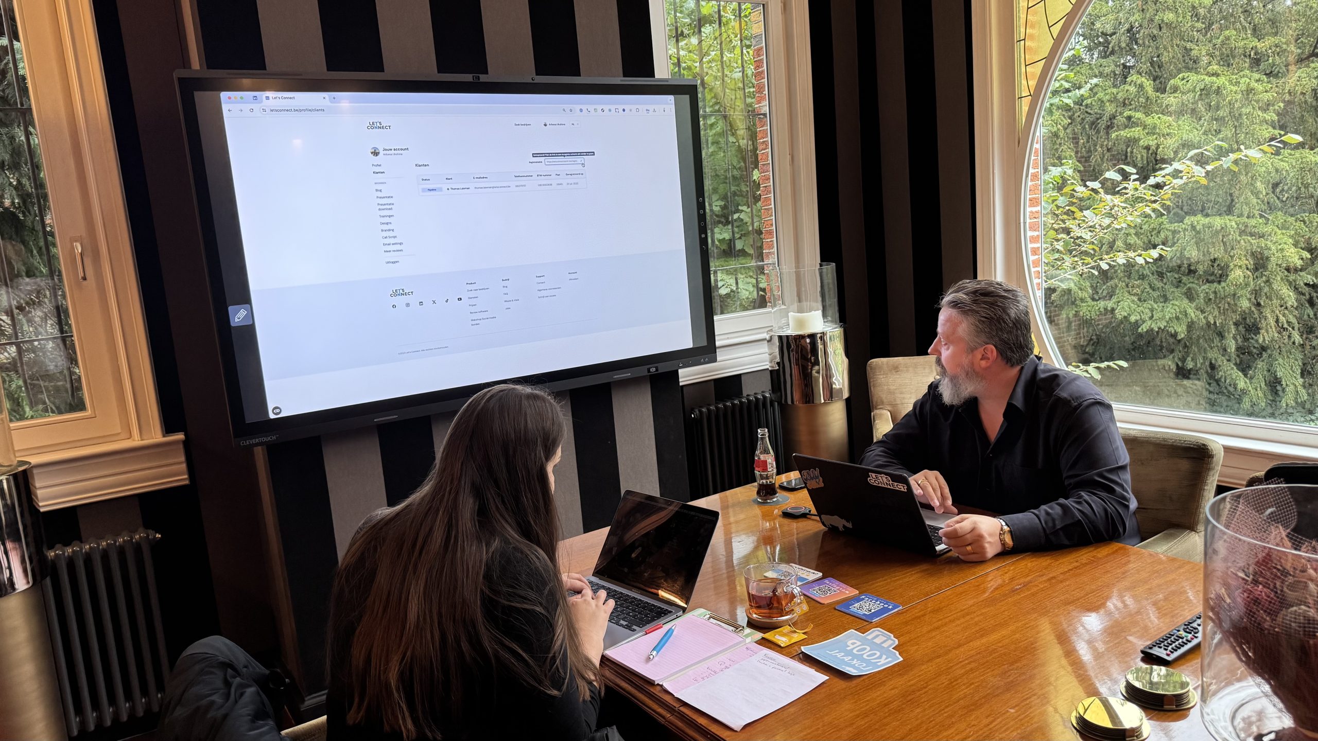 oplei Twee mensen werken aan laptops tijdens een vergadering, met een groot scherm en ramen met uitzicht op bomen.